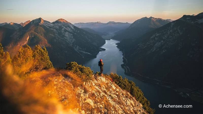Wanderer steht bei Sonnenaufgang am Grad und blickt auf den Achensee